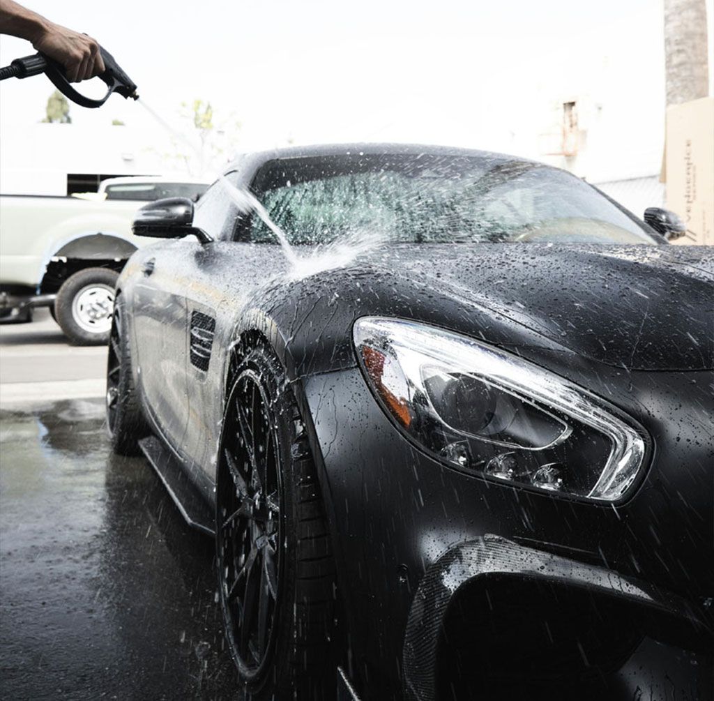 Black sports car being washed with a hose, covered in soap suds outdoors