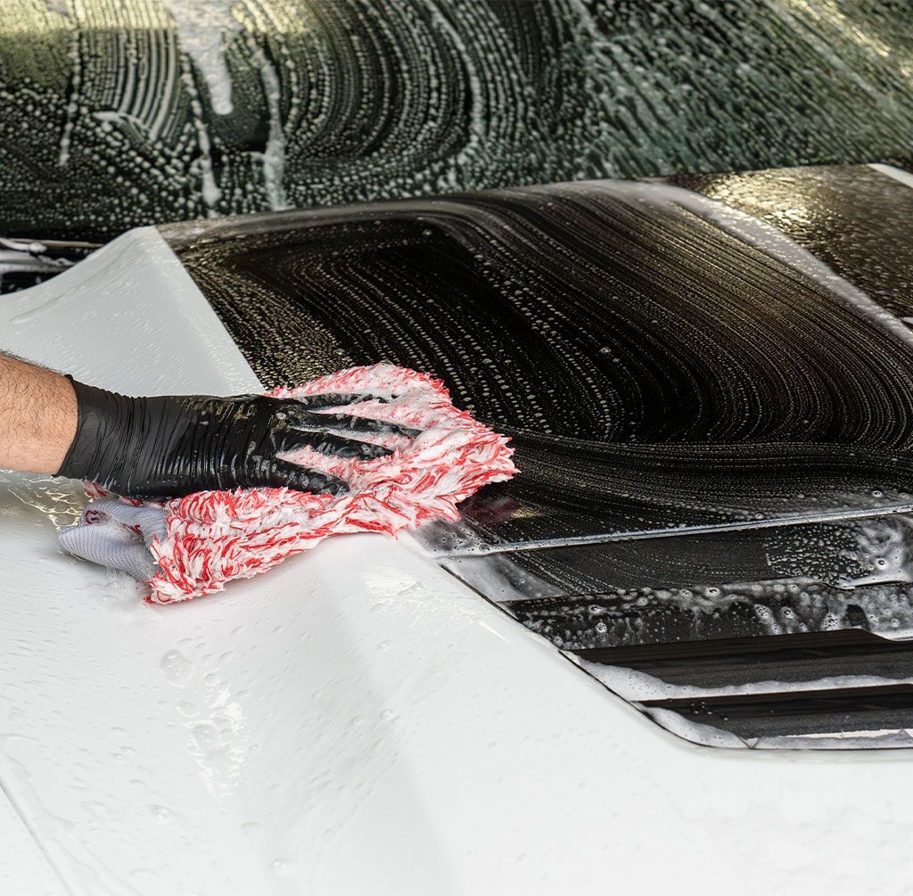 Hand washing a black car hood with a red-and-white sponge and black glove