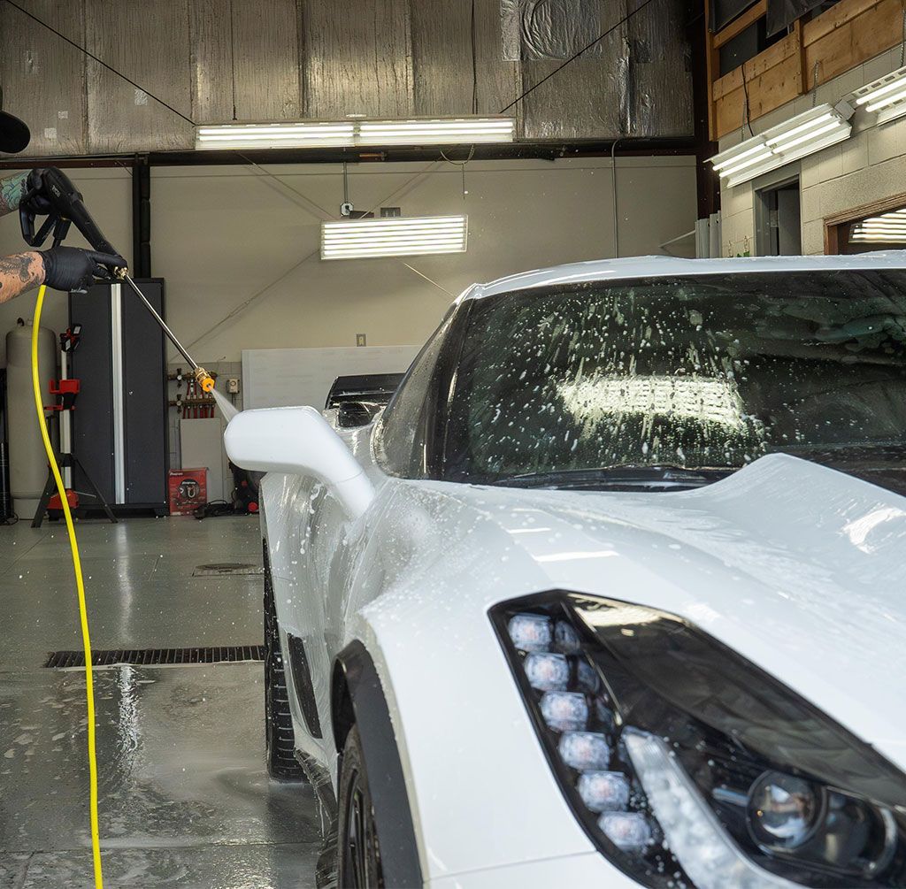 White sports car being washed in an indoor car wash, with water spraying across the hood and windshield.