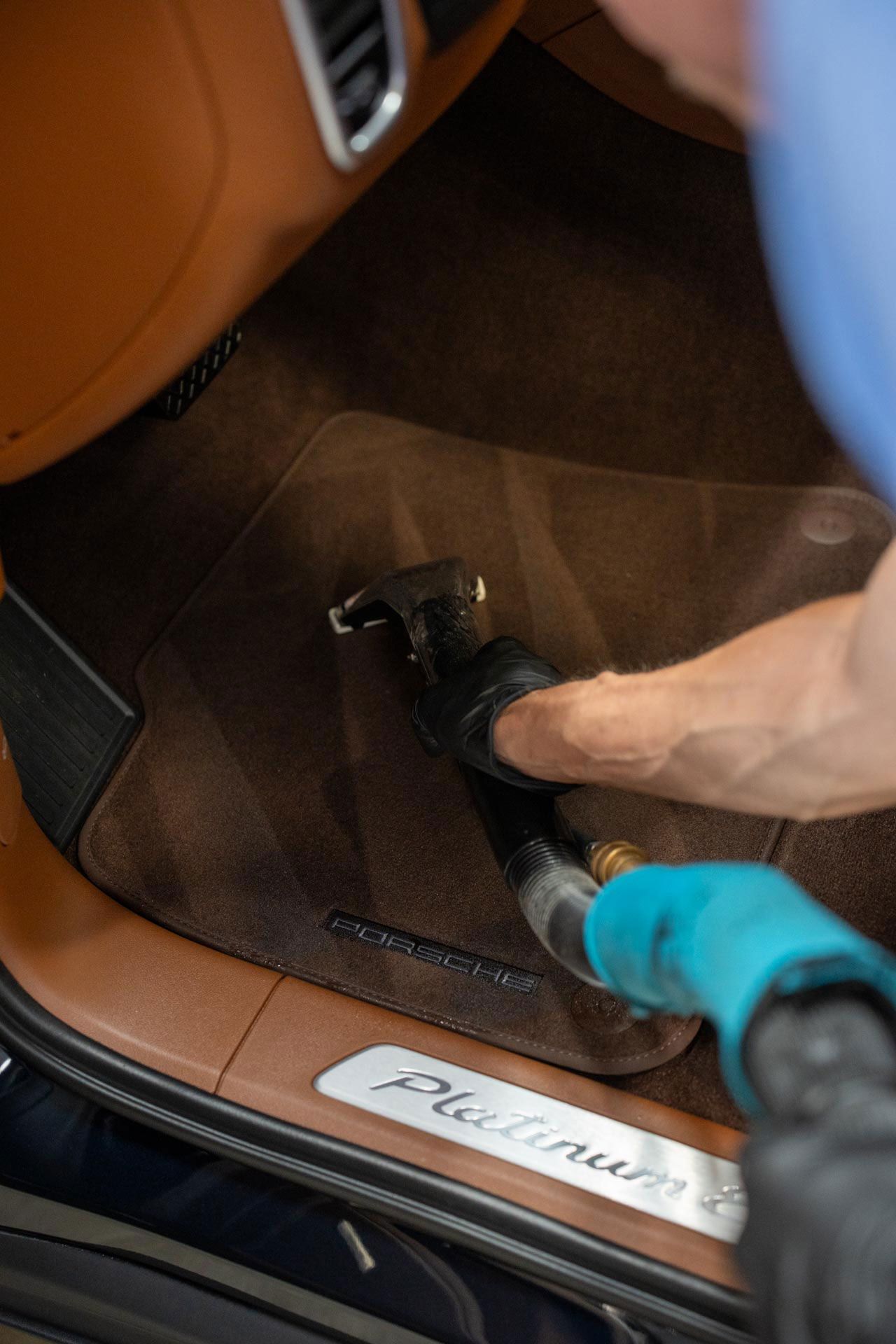 Person fastening a black sandal in a brown car footwell near a door sill labeled “Porsche”