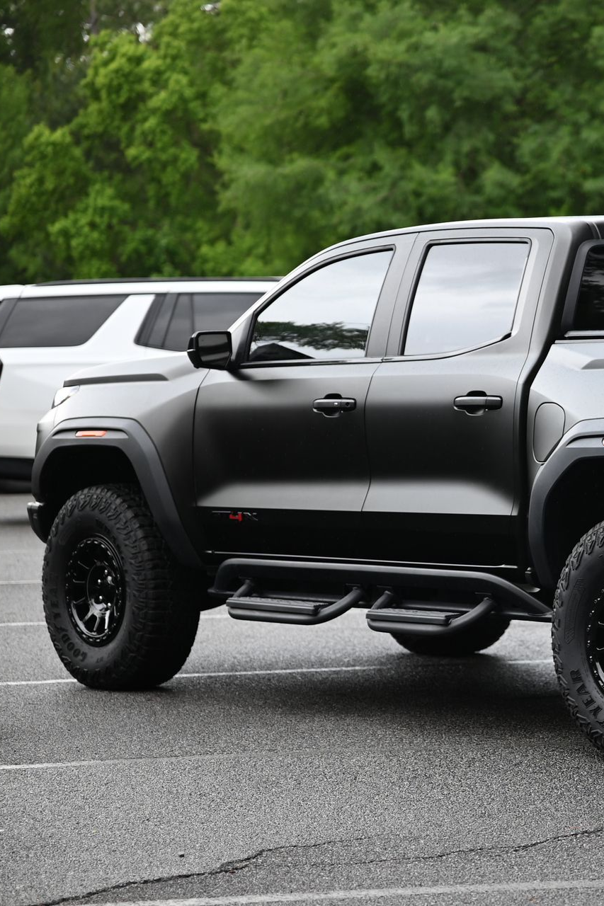 Black pickup truck parked on wet pavement, side view, with green trees in the background