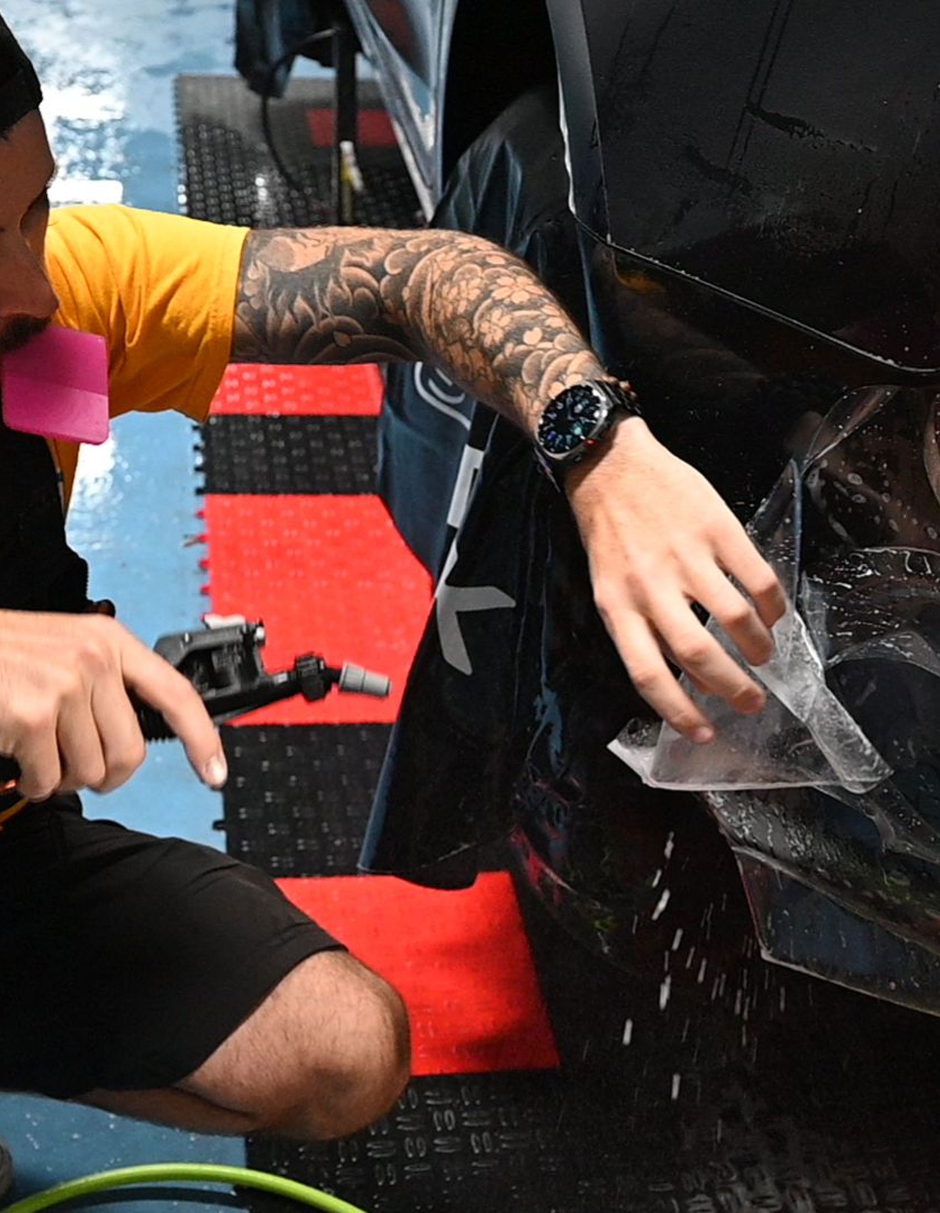 Person in tattooed arm and black watch kneels beside a black panel, peeling back clear plastic wrap.