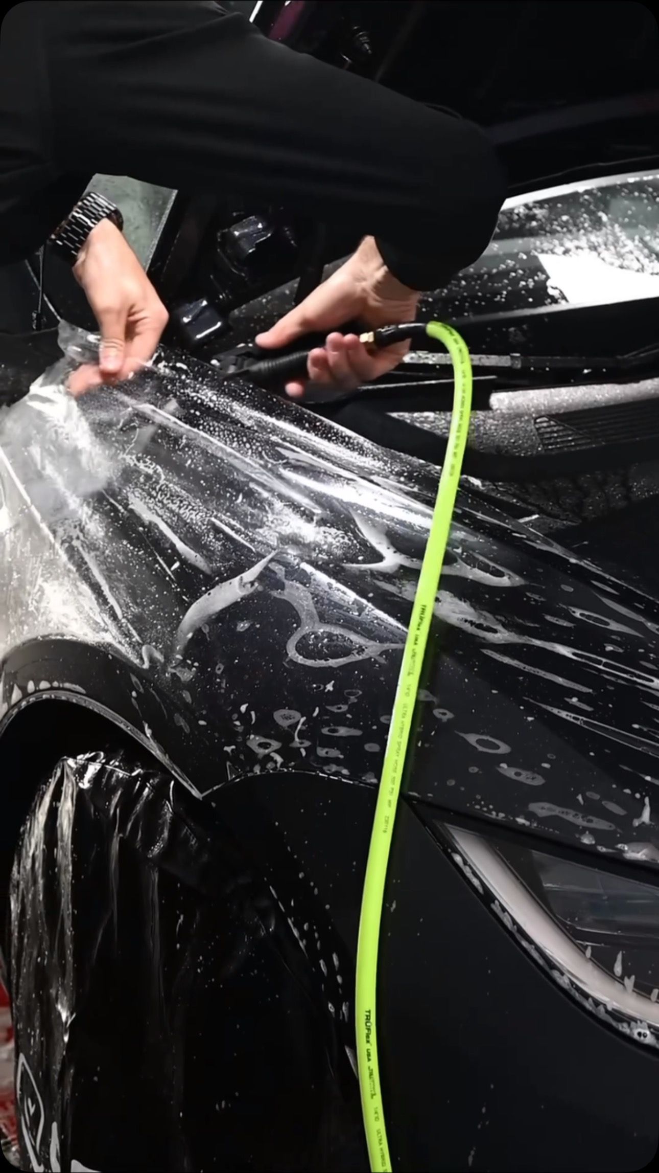 Hands using a hose to wash a black car covered in soap suds