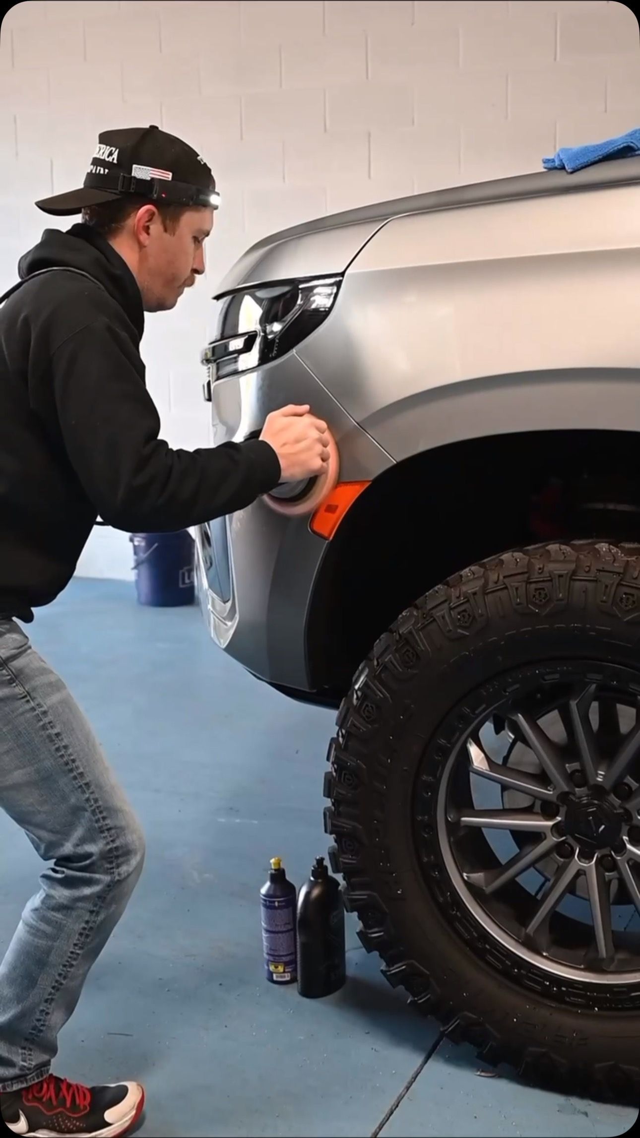 Person cleaning the front fender of a silver SUV in a garage, with a bottle on the floor.