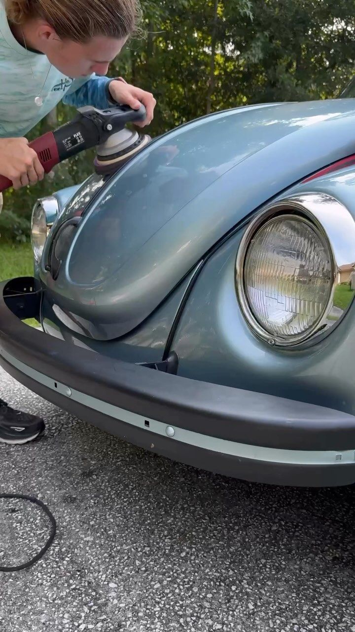 Person polishing the hood of a blue-gray classic Volkswagen Beetle with a buffer outdoors