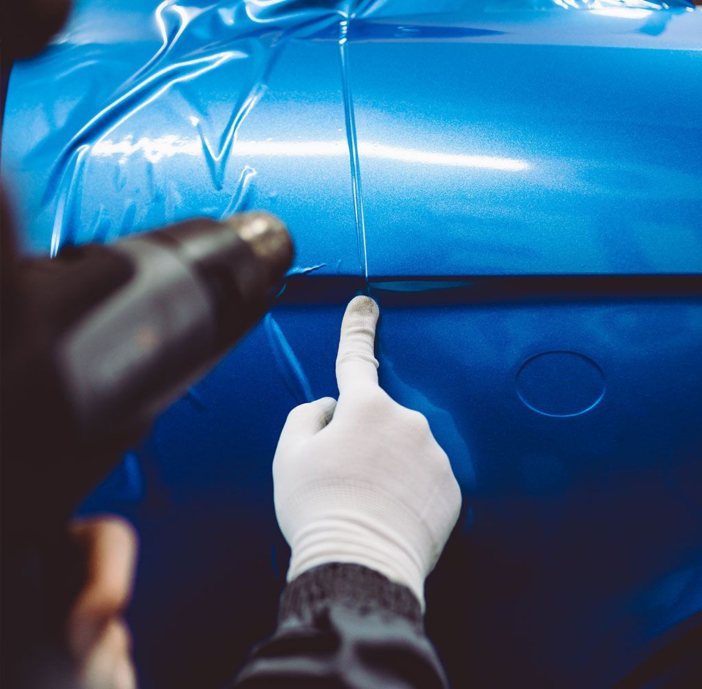 Gloved hands applying blue protective film to a car body with a heat gun