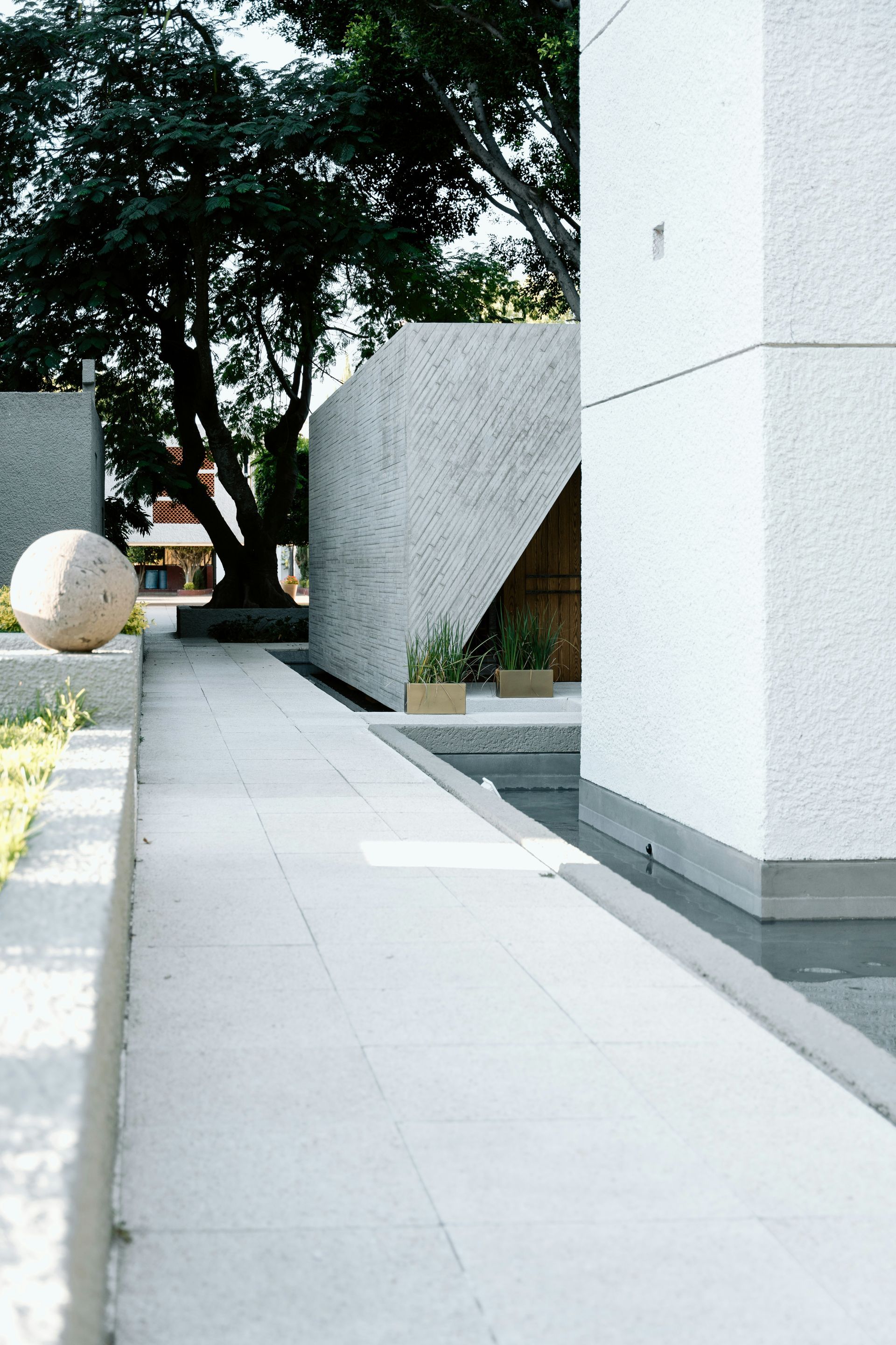 Outdoor walkway beside white concrete buildings and trees, with a large round stone sculpture.