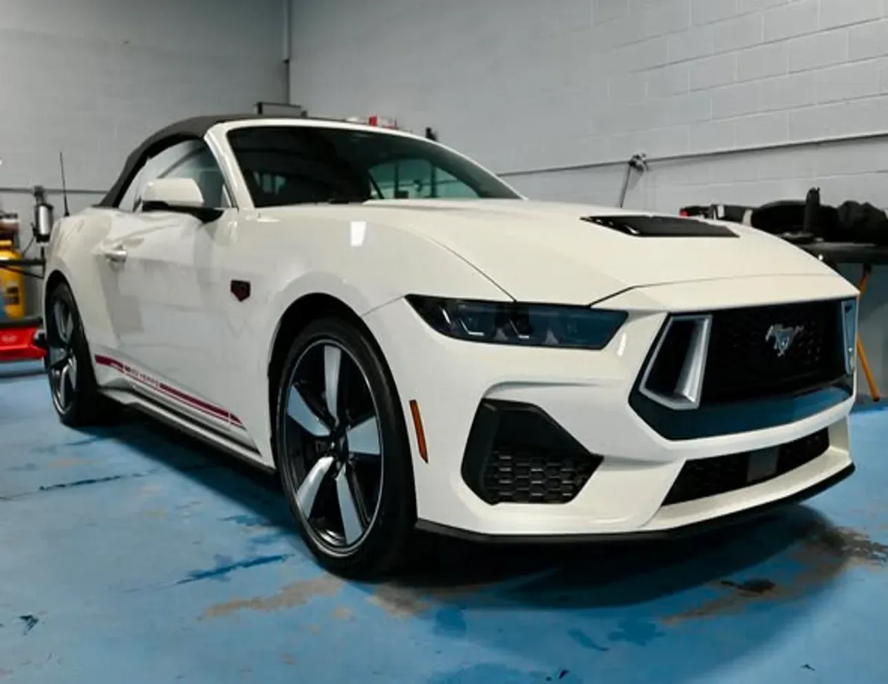 White Ford Mustang in a garage, angled front view on a blue floor.
