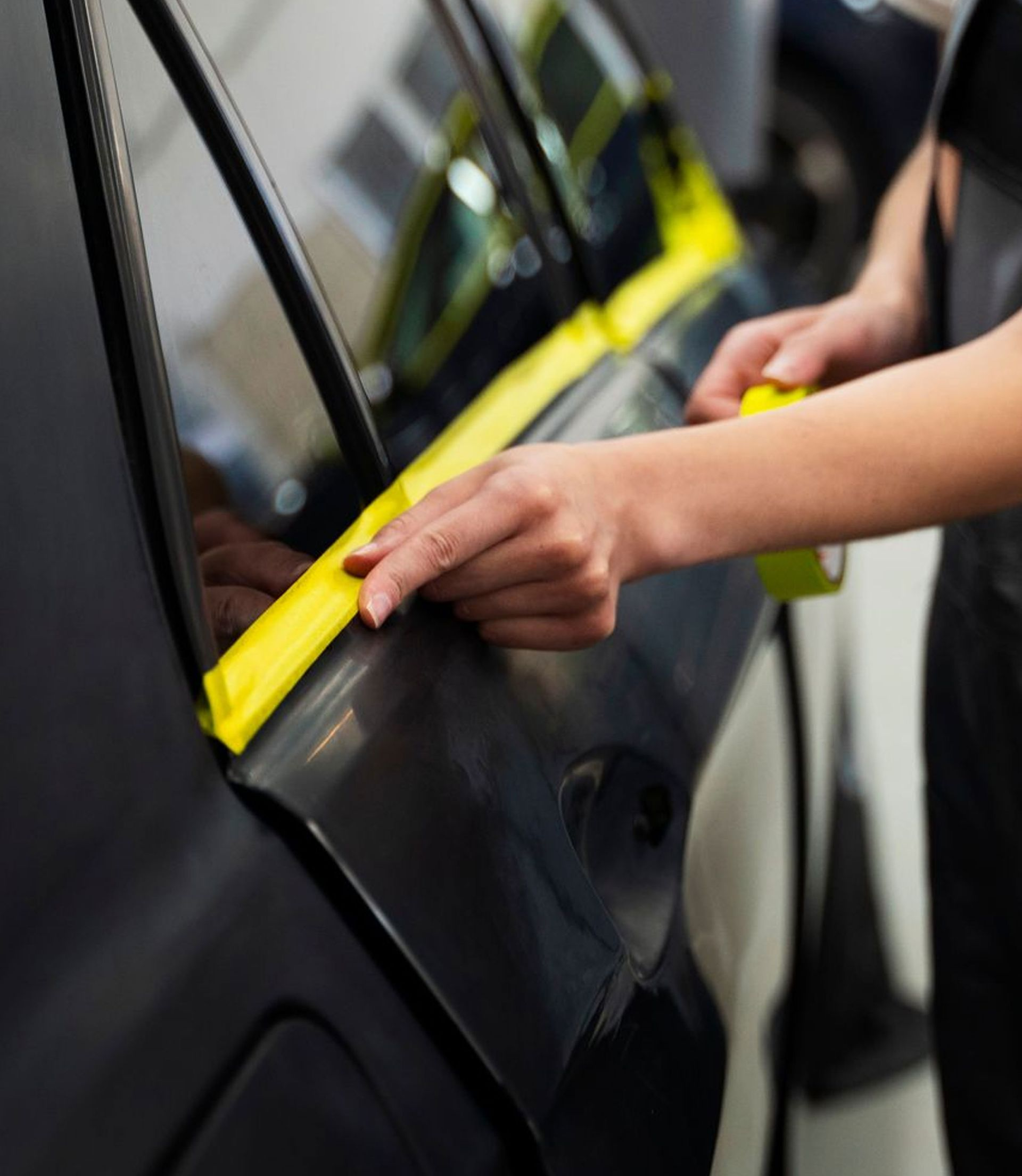 Hand placing yellow tape on a black car door at an auto repair shop