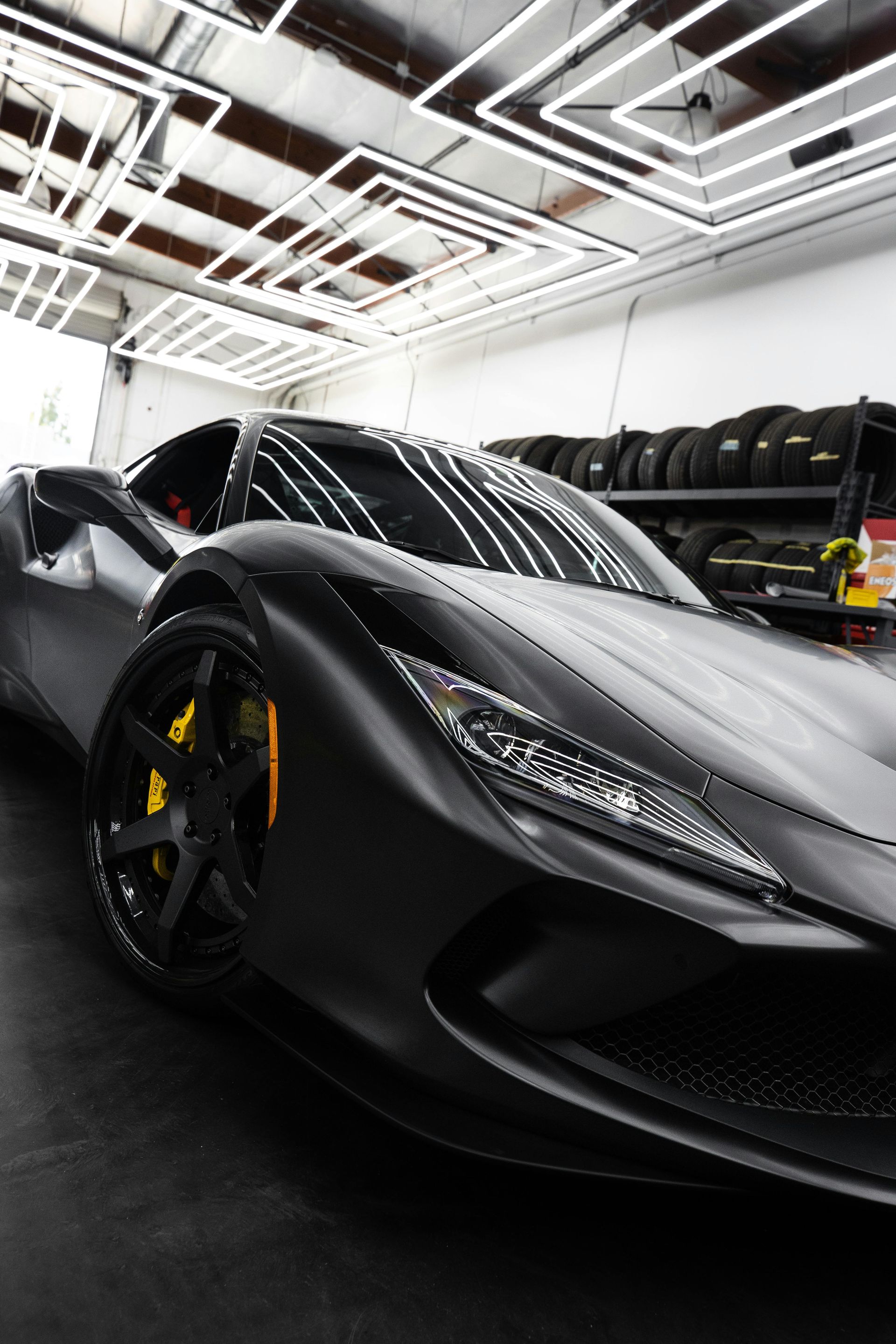 Black sports car in a modern garage with bright ceiling lights and stacked tires in the background