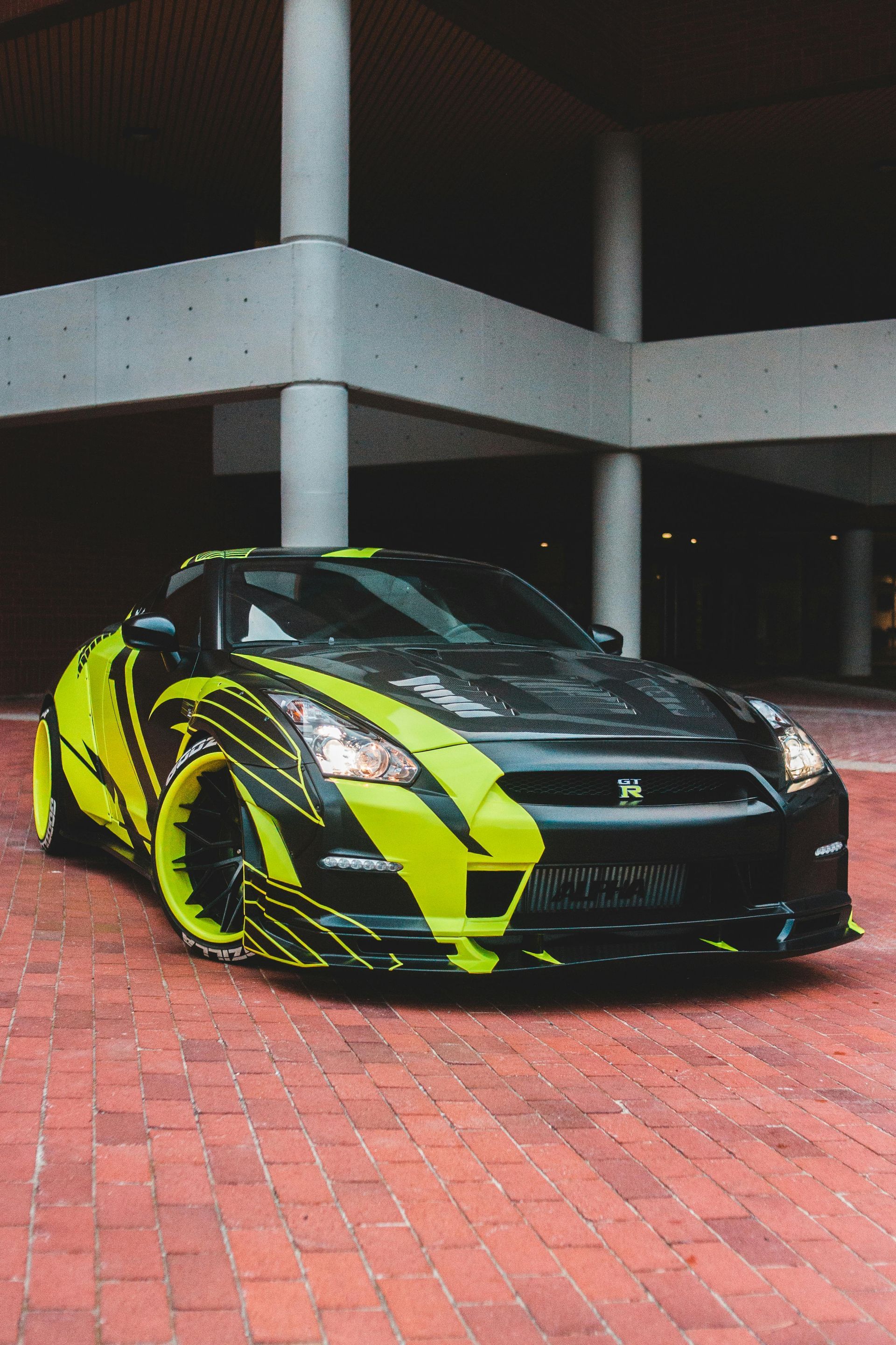 Black sports car with neon yellow accents parked on a red brick driveway under a concrete structure