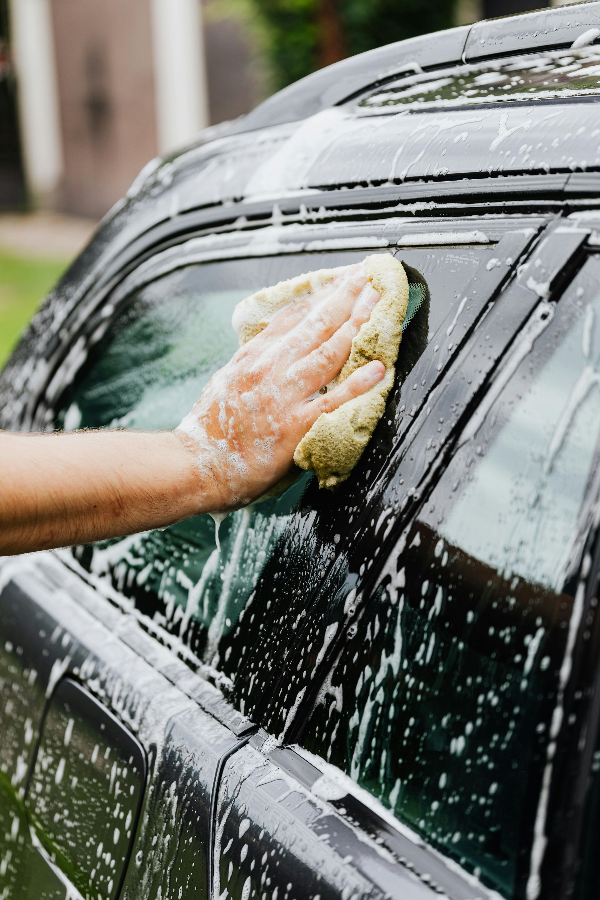 Hand washing a black car with a soapy sponge, suds dripping down the window
