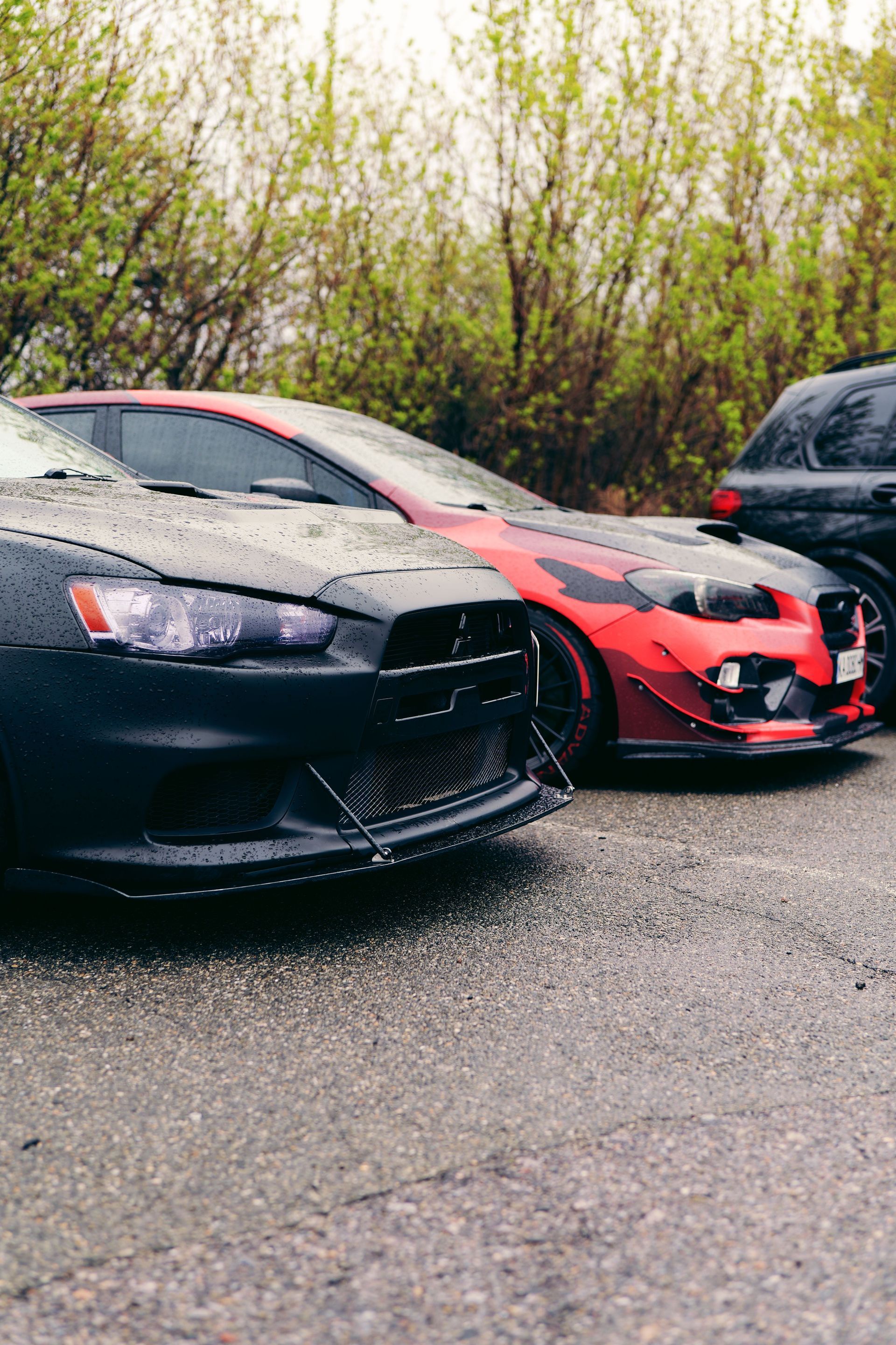 Two sports cars, one black and one red, parked on a wet road by dense bushes