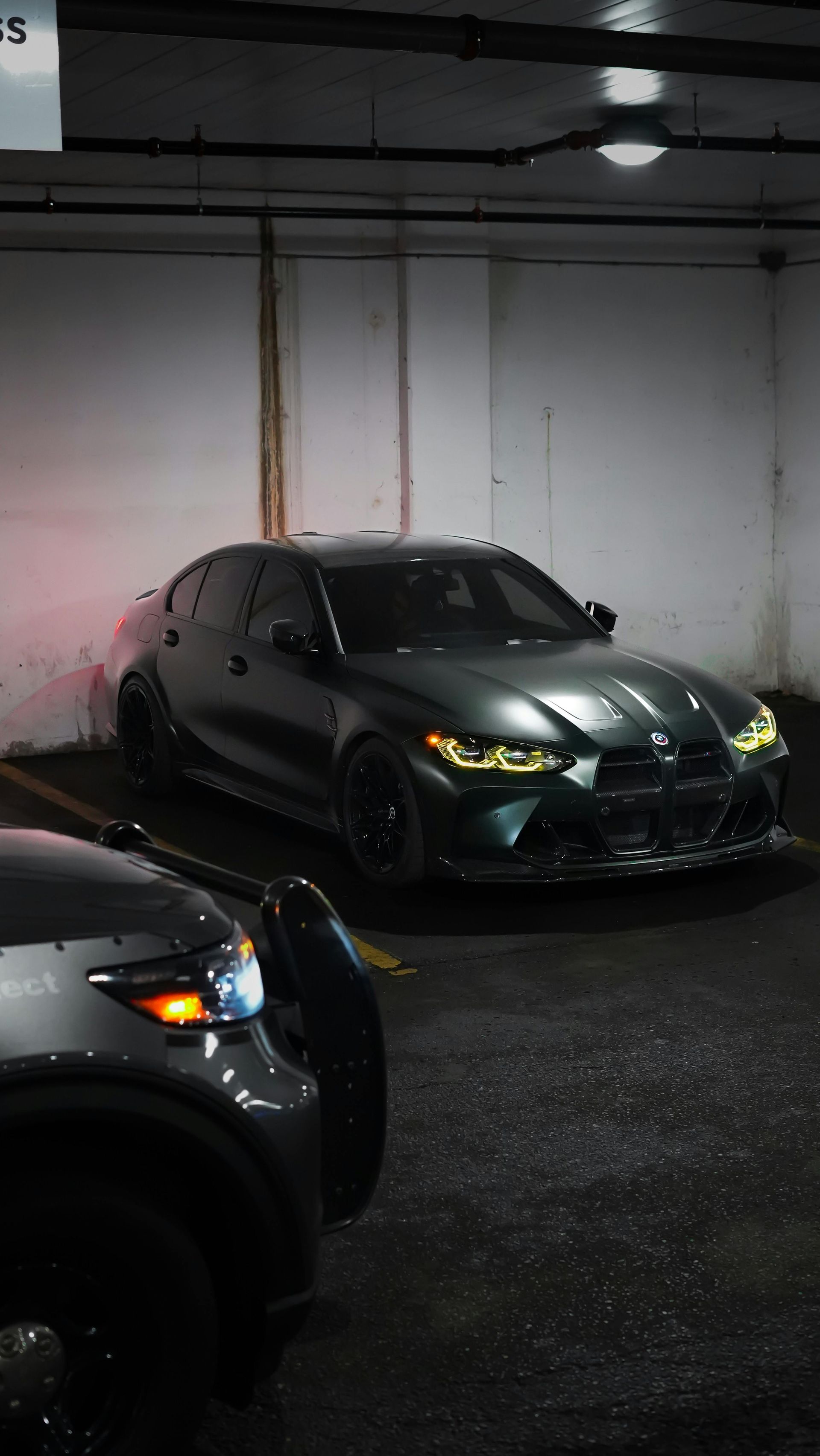 Two dark sports cars parked in a dim garage, one centered with green headlights on