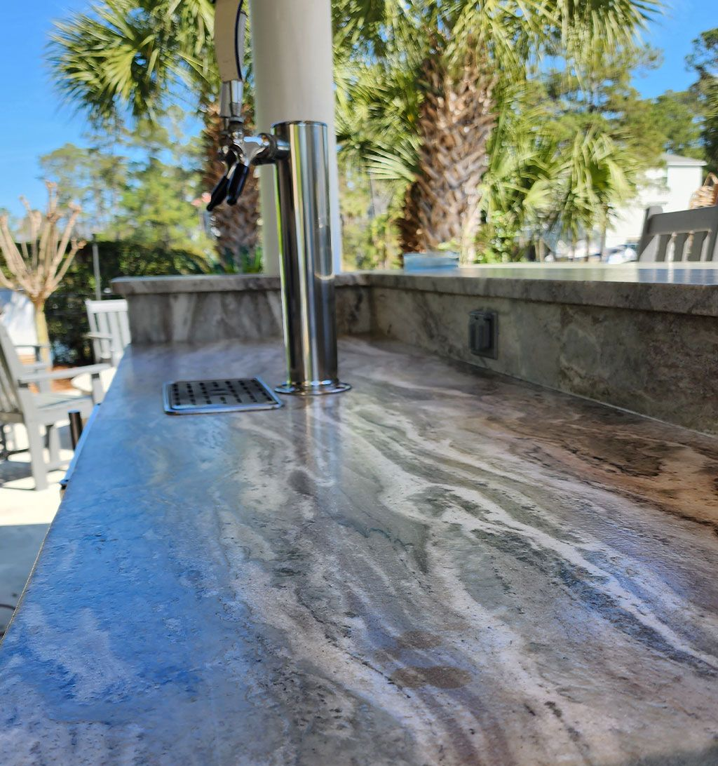 Outdoor stone counter with a chrome water faucet and drain, wet from running water, beside palm trees and a pool.