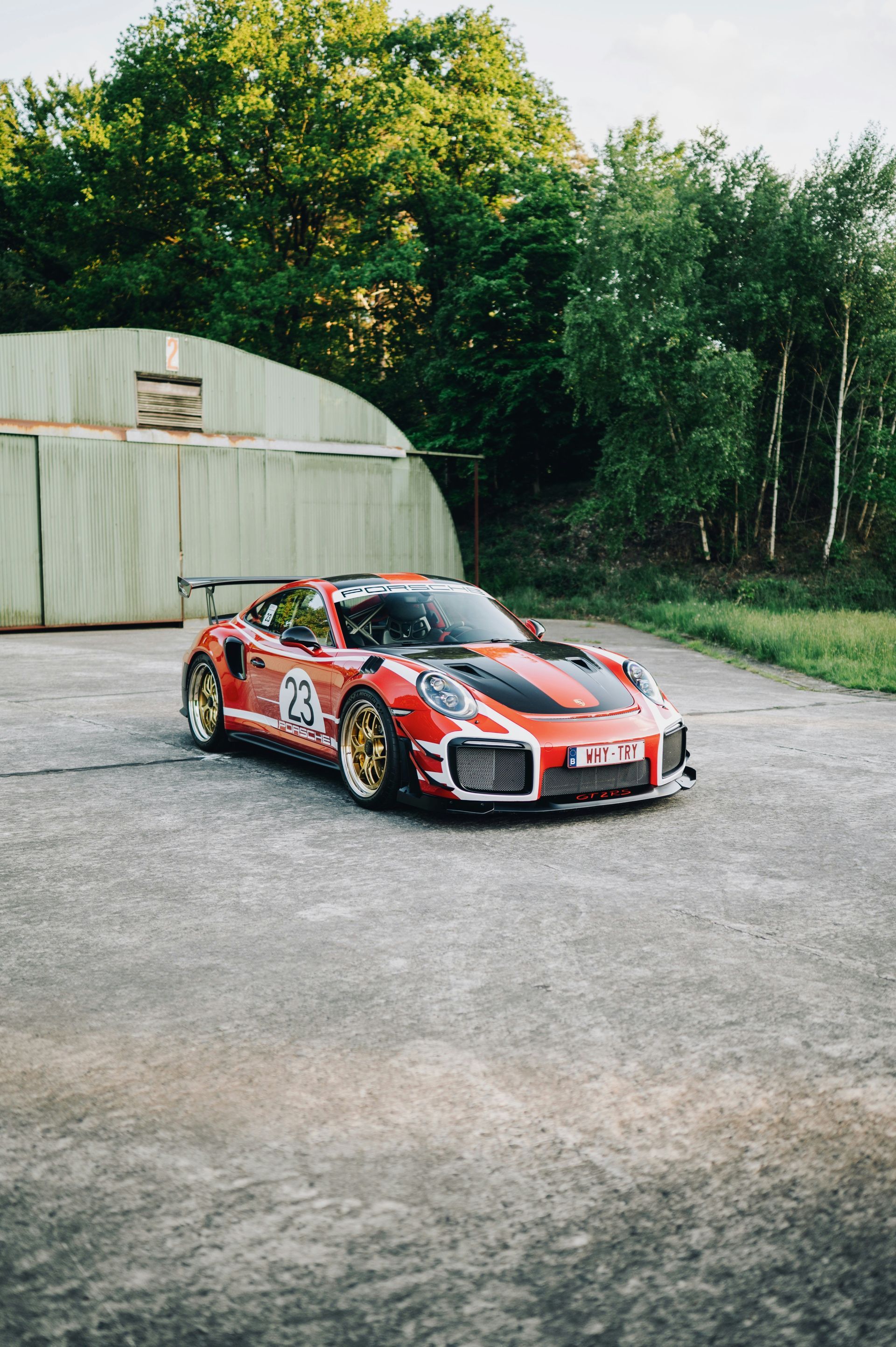Red and black race car parked on gravel beside a greenhouse and trees