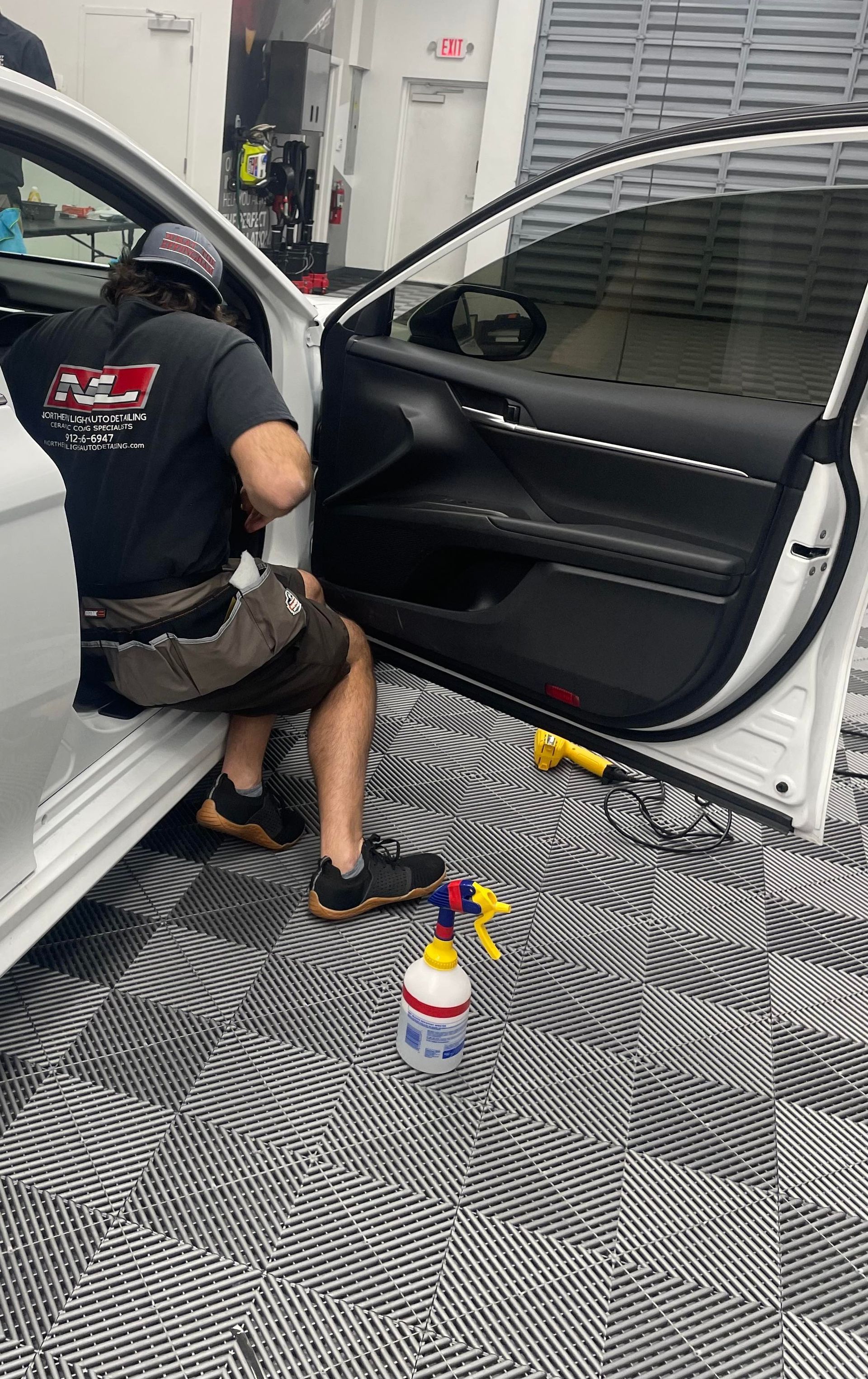 Person detailing a white car interior with a spray bottle and tools on the floor in a garage-like space