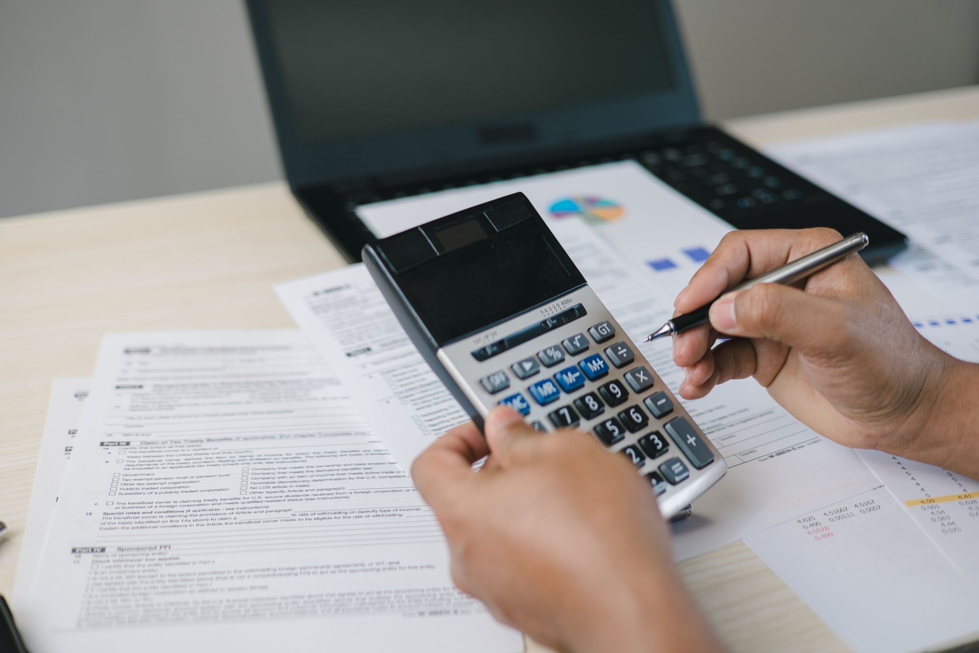 Hands using a calculator beside paperwork on a desk, with a laptop in the background.