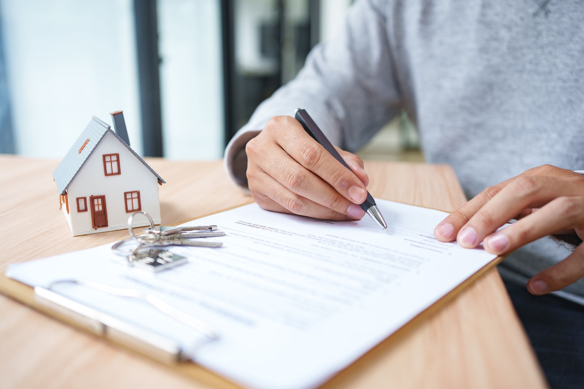 Hands signing a document beside a small house model and keys on a desk