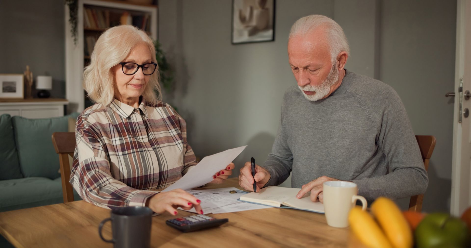 Two people reviewing documents at a kitchen table, one writing while the other points at paperwork.