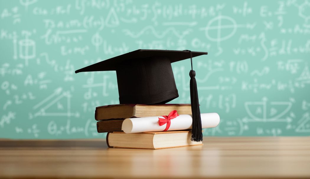 Graduation cap on stacked books with a diploma in front of a chalkboard