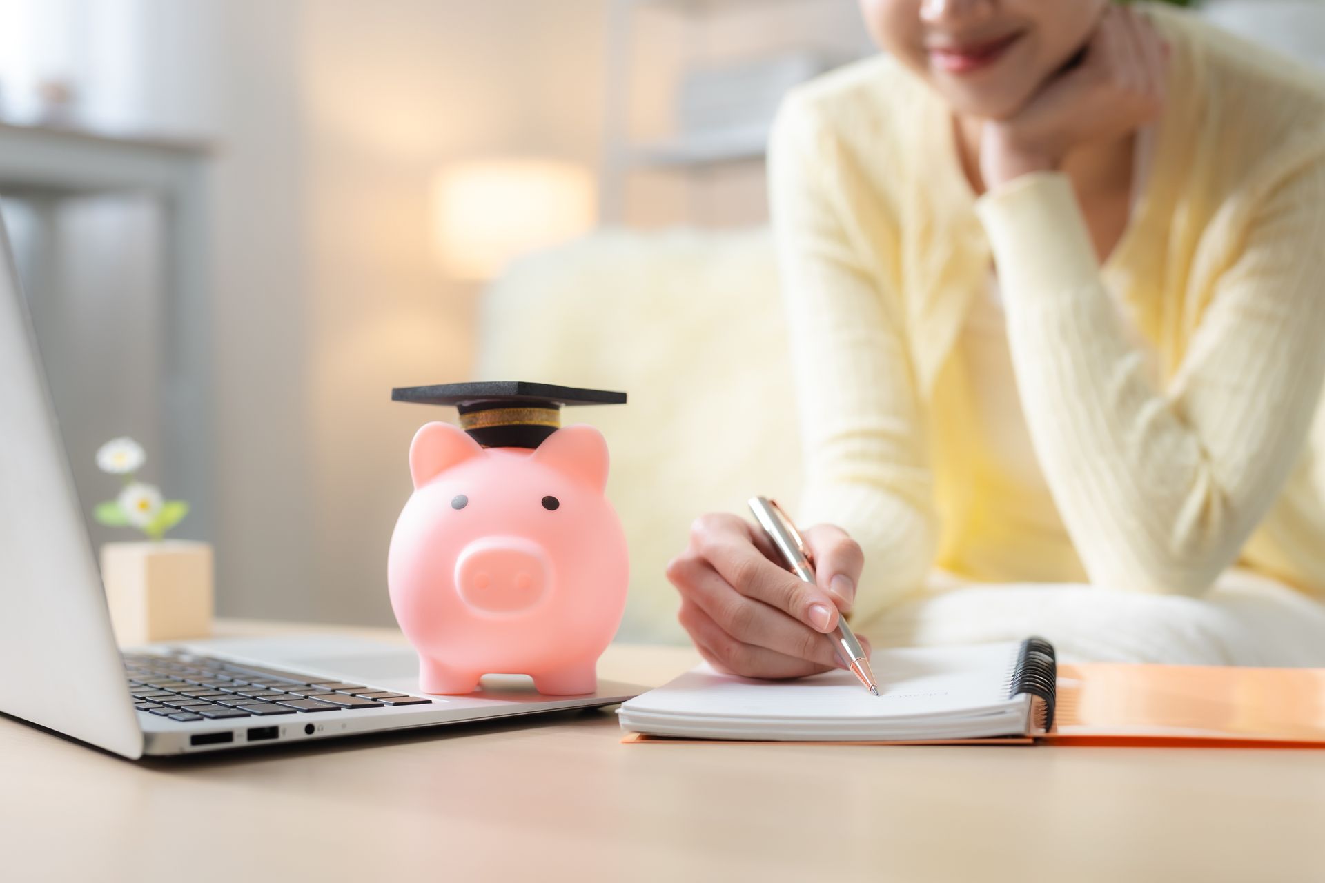 Person studying at a desk with laptop, piggy bank, notebook, and pen in a bright room