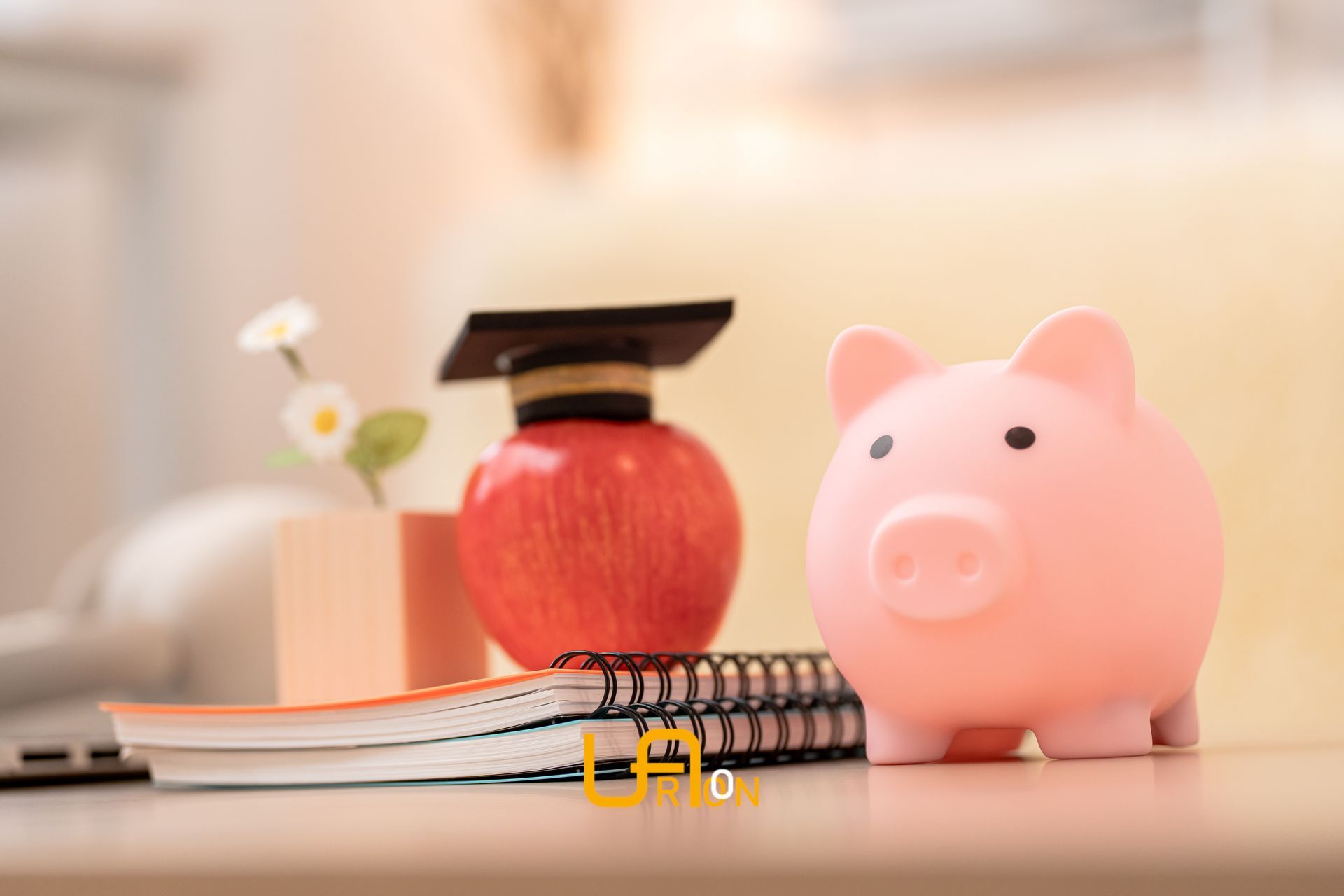 Pink piggy bank beside red apple, notebook, and small white flowers on a desk