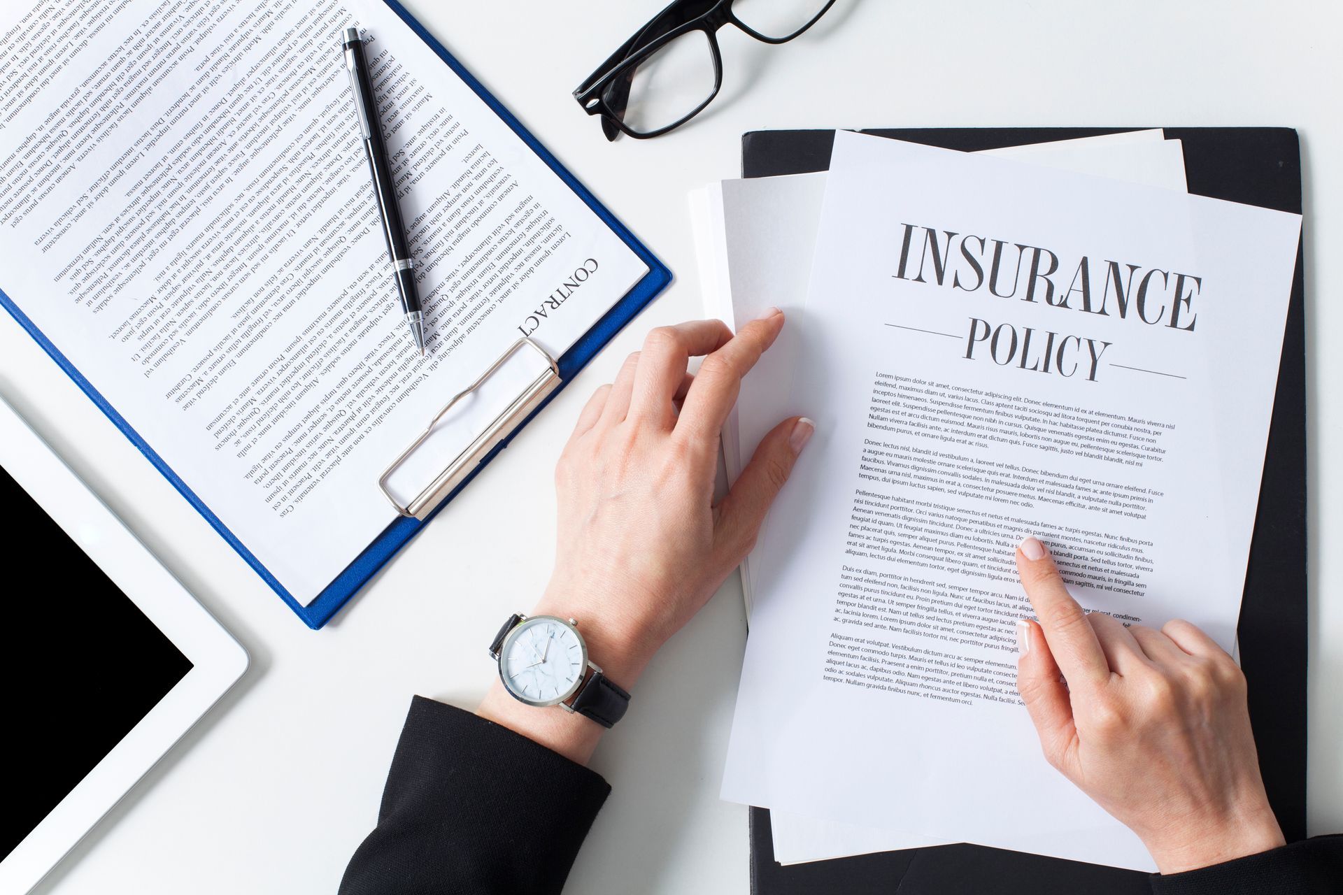 Hands reviewing an insurance policy document beside a tablet, glasses, and an open notebook on a desk
