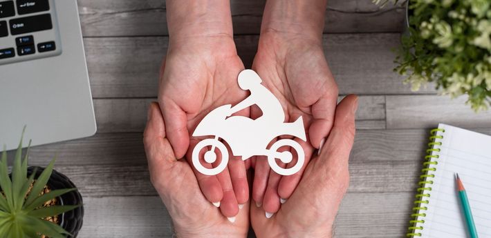 Hands holding a white motorcycle icon above a wooden desk with a laptop, plant, and notebook
