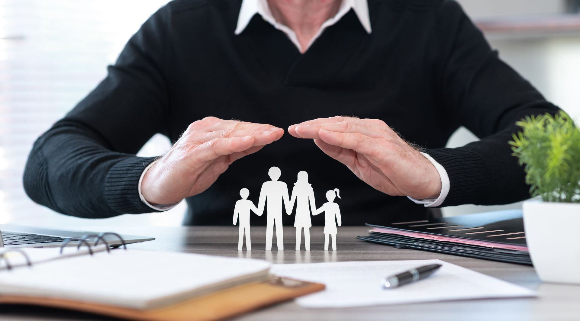 Businessperson shielding small family figures on desk, symbolizing insurance or protection