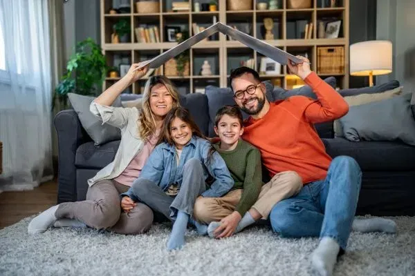 Family sitting on a living room rug, smiling and holding a blanket roof together.