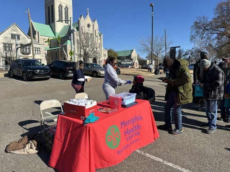 People gathered at a Memphis Health Center table with a red cloth on a sunny street near a church.