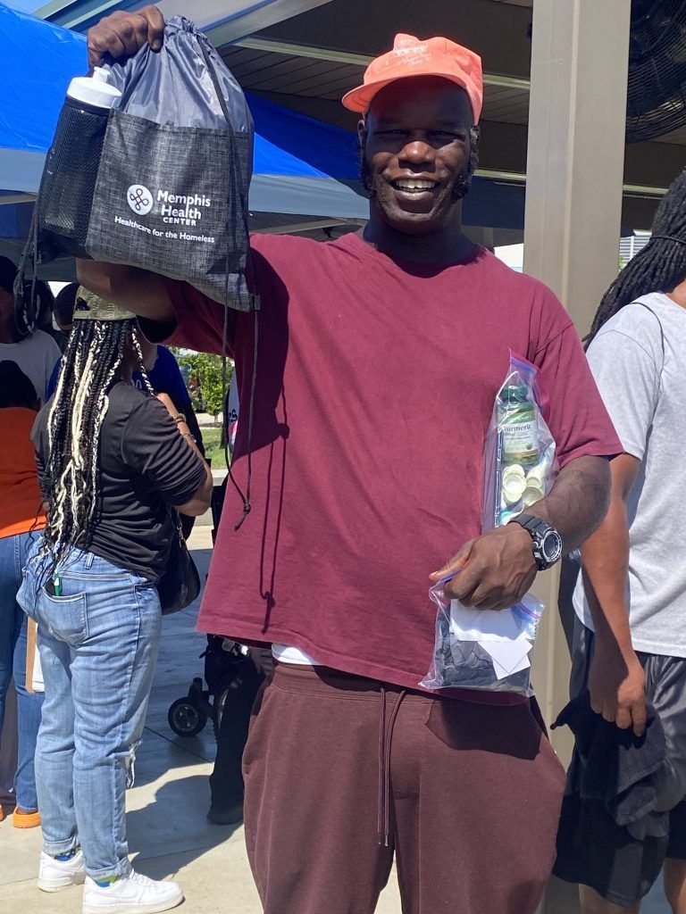 Man smiling and holding up a black bag outdoors, with a woman and another person in the background.