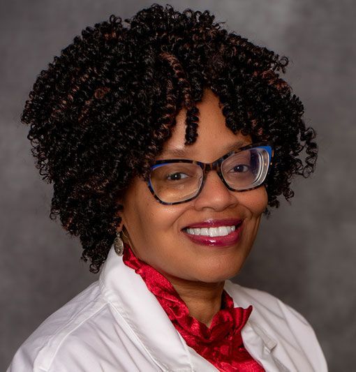 Studio portrait of a smiling woman wearing glasses, a white jacket, and a red scarf against a gray background