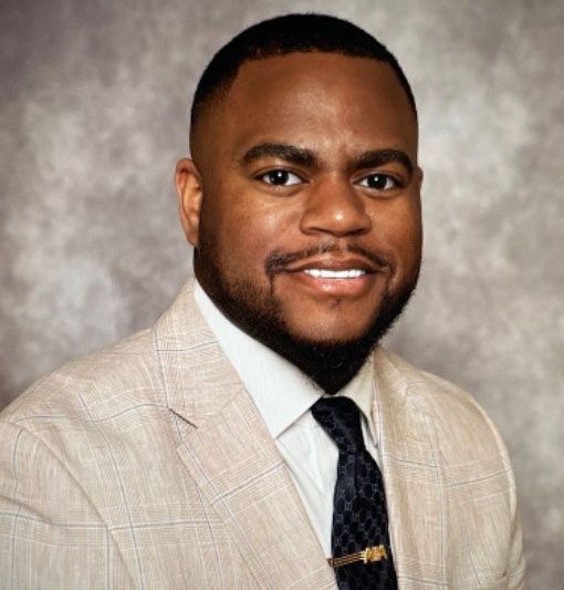 Smiling man in a light tan suit and dark tie against a gray studio backdrop