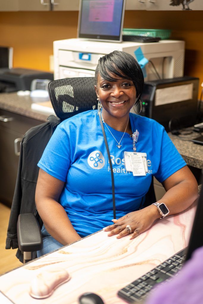 Smiling employee in blue shirt seated at a desk in an office, with computer and printer behind her.