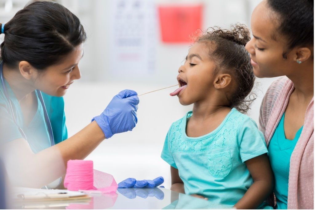 Medical professional swabbing a child’s throat while a caregiver holds her in a clinic