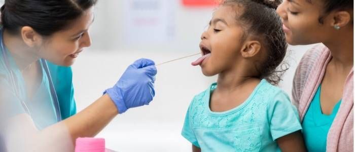 Health worker swabbing a child’s mouth while another adult supports her in a clinic