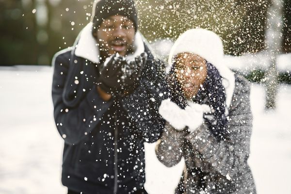 Two people in winter coats catch snowflakes outdoors, smiling in a snowy park.
