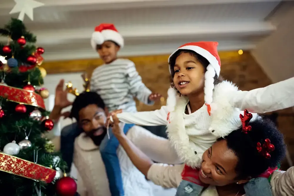 Family in Santa hats decorating a Christmas tree, with children smiling and playing indoors