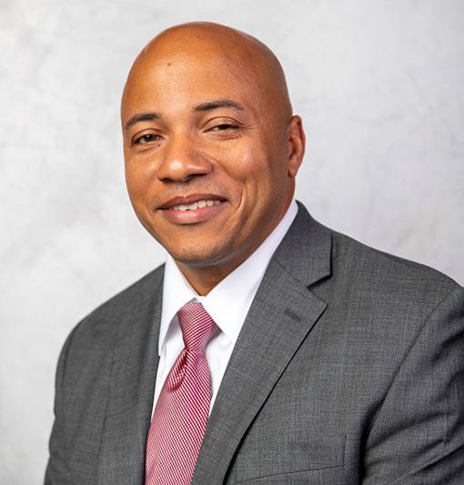 Professional headshot of a smiling person in a dark blazer and blue shirt against a blue backdrop
