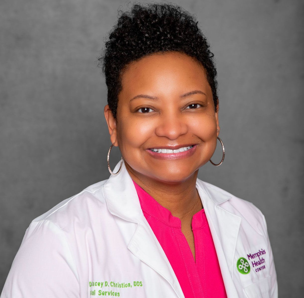 Smiling healthcare professional in a white coat and pink blouse against a gray background