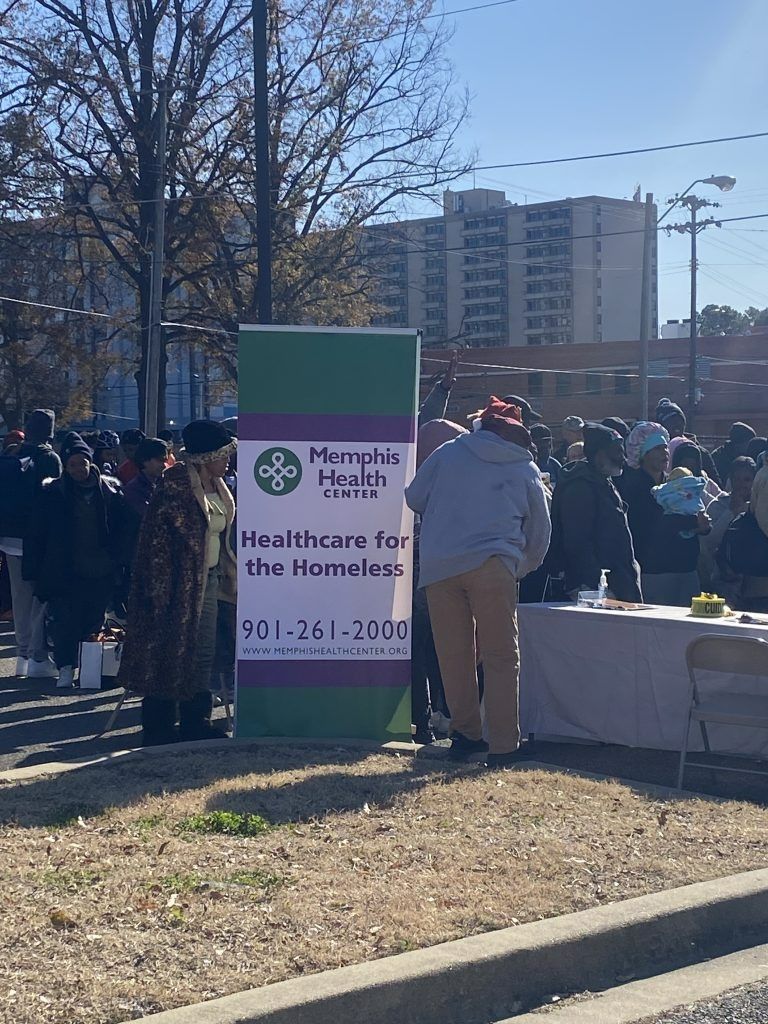 Outdoor community event with a Hampton Health sign, people gathered, and a table in a park.