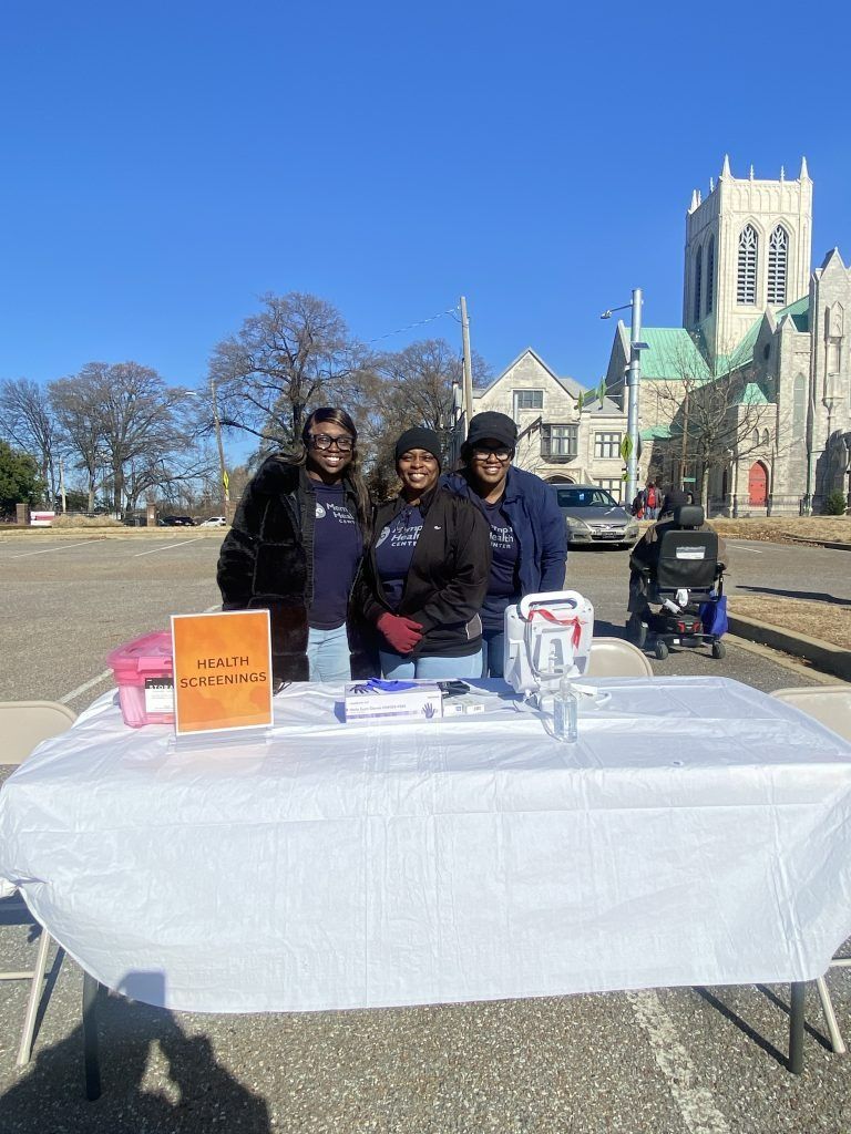 Three people behind a white table outdoors, with signs and a church-like building in the background.