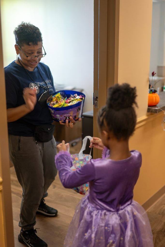 Adult offering candy to child in purple dress in a hallway, both smiling and facing each other