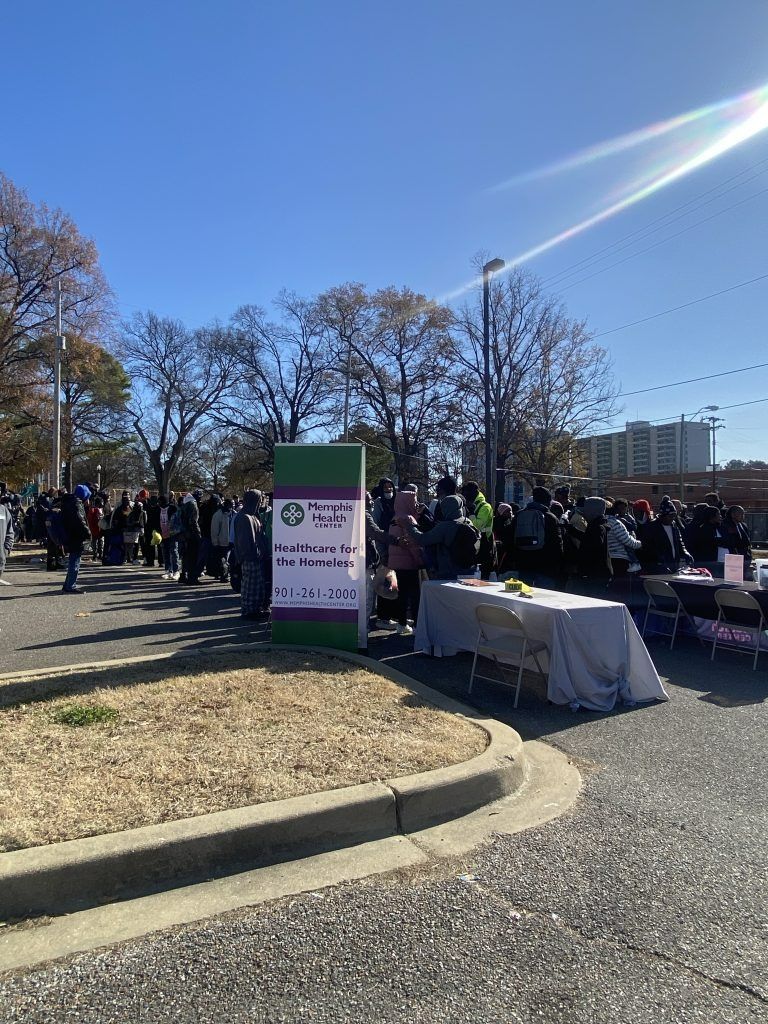 Crowd gathered outdoors near a sign and table on a sunny day