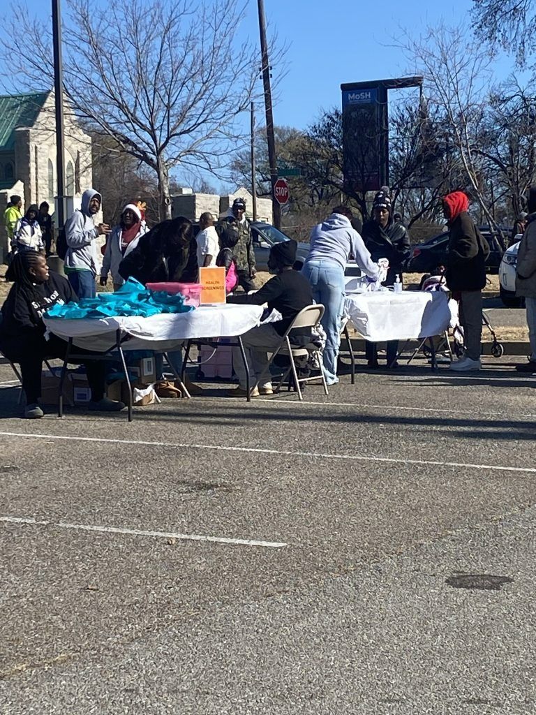 People gathered around tables outdoors on a sunny day, with bags and supplies in a parking lot.
