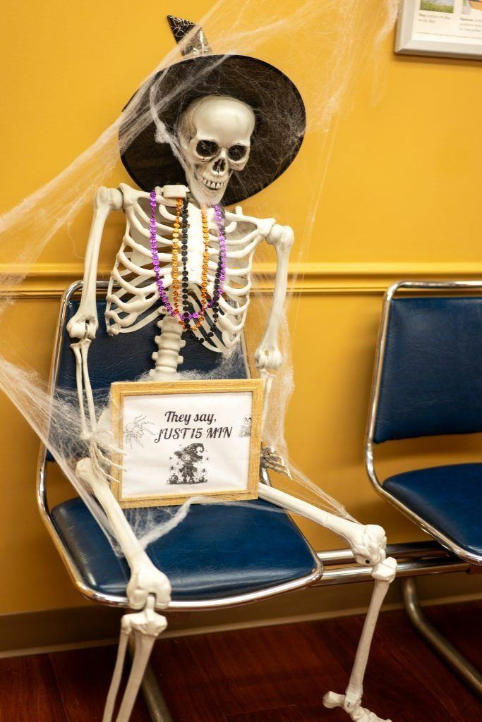 Skeleton mannequin in a black hat, wrapped in cobwebs, seated on a blue chair with a sign in front.