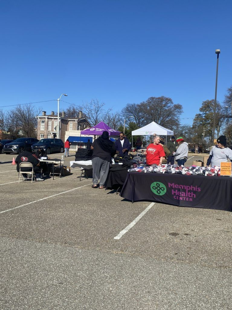 Outdoor community market with vendor tents, people browsing, and a table of items in a parking lot under a blue sky