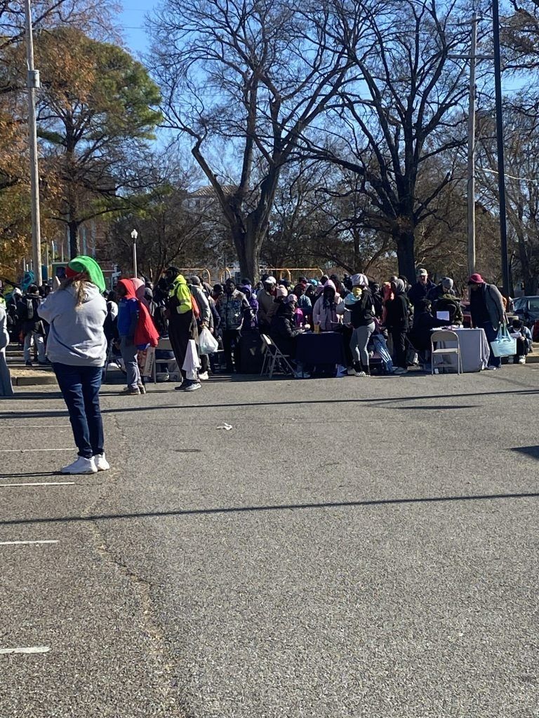 Crowd gathered outdoors near tables in a park on a sunny day, with bare trees and people in jackets.